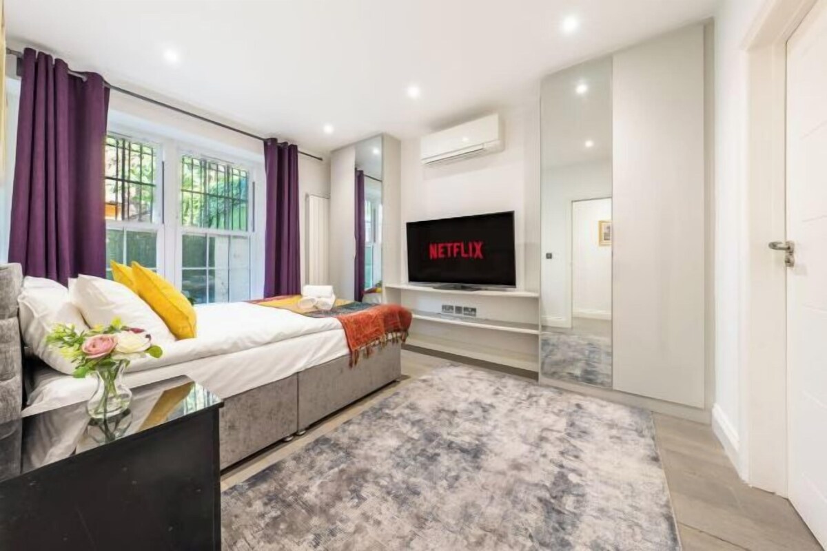 Un dormitorio moderno con cama, televisión y ventana con cortinas moradas.