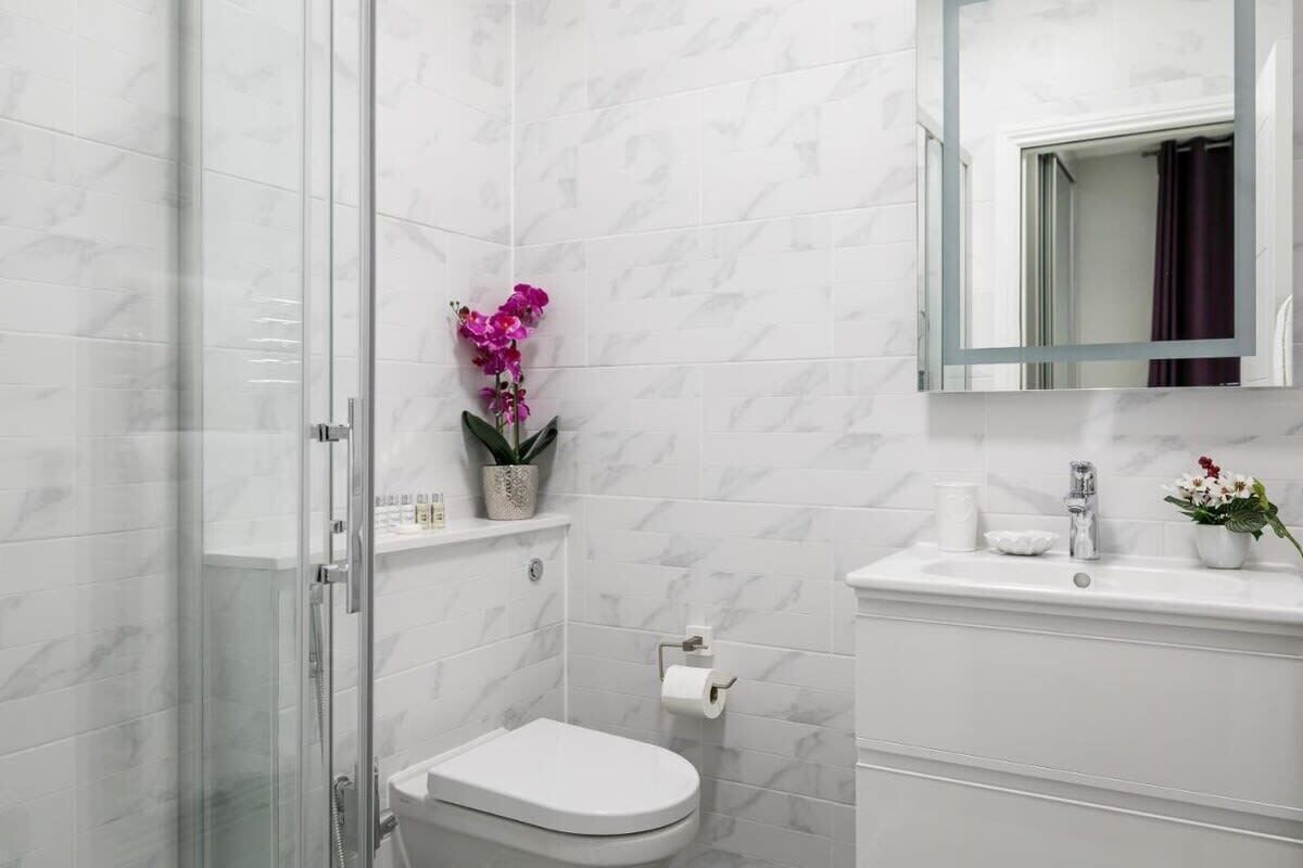 A modern bathroom with a glass shower enclosure, a white toilet, a marble backsplash, a sink with a mirror above it, and a small shelf with a potted plant.