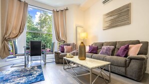 A living room with a sofa, a coffee table, a lamp, and a chair. There is a window with curtains and a view of greenery outside. - Maida Vale - Carlton Vale Apartments (London)