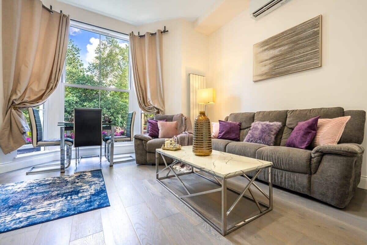 A living room with a sofa, a coffee table, a lamp, and a chair. There is a window with curtains and a view of greenery outside.