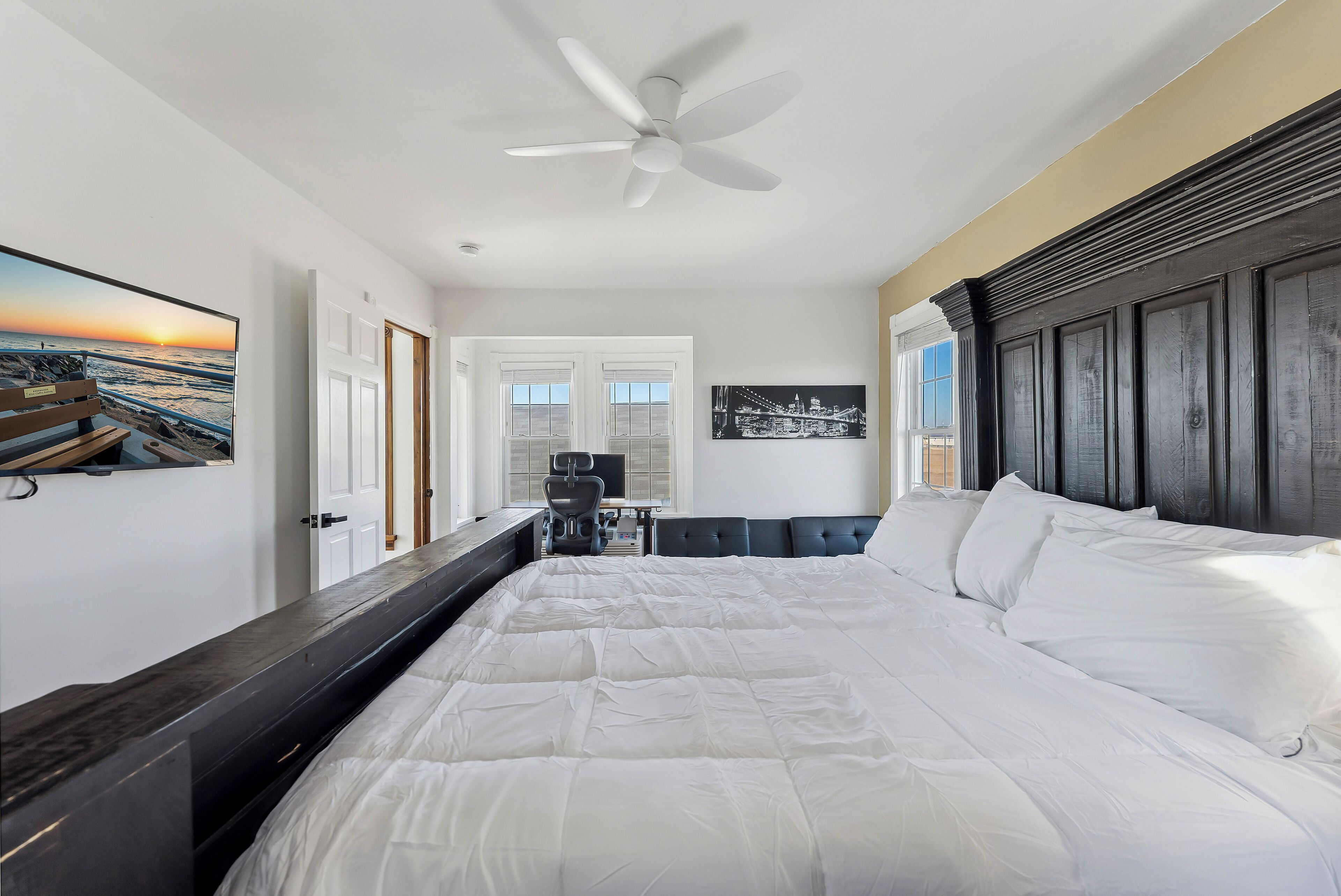 A hotel room with a large bed, a ceiling fan, a desk with a chair, and a view of the ocean through the window.