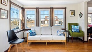 Ein Wohnzimmer mit einem weißen Sofa, einem grünen Sessel und einem schwarzen Stuhl. Große Fenster mit Holzrahmen bieten Blick auf umliegende Gebäude.