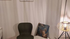 Una habitación con un sillón verde oscuro, un cojín estampado con flores, una lámpara de pie y un cuadro en la pared.