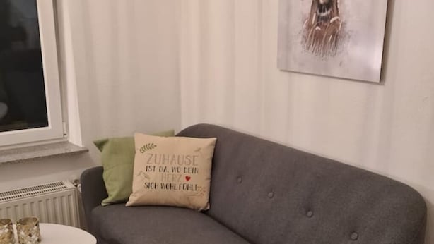 Un sofá gris con un cojín que tiene texto en alemán, una mesa blanca con dos vasos, y un radiador debajo de la ventana.