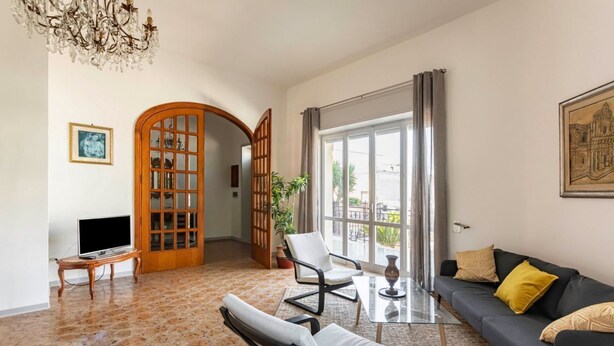 A living room with a sofa, chairs, a TV, a chandelier, a large window, and a door leading to another room.