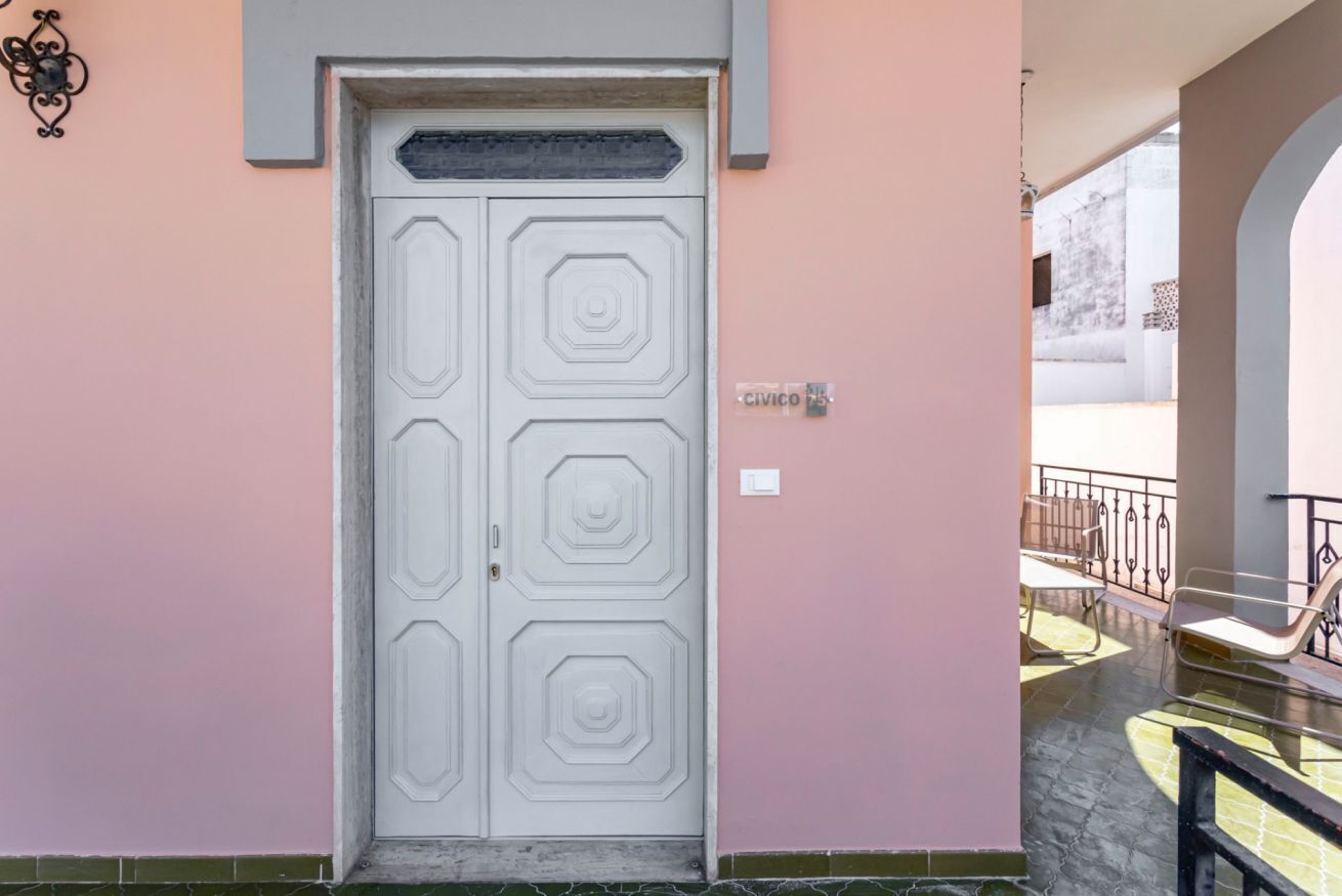 A white door with a geometric pattern, a pink wall, and a balcony with chairs.