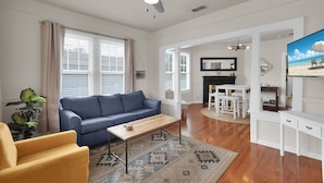 A spacious living room with a blue sofa, a wooden coffee table, and a yellow armchair. There is a ceiling fan, a fireplace, and a kitchen area visible through an open doorway. - Coastal Cottage near Winery (St. Augustine)