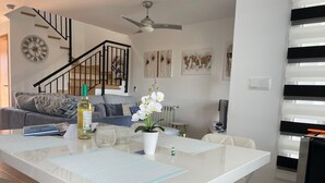 A modern kitchen with a central island, a dining area with white chairs, a staircase, and artwork on the walls.
