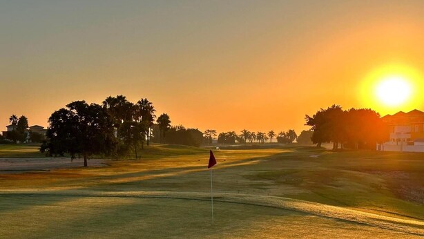 A golf course at sunset with a flag in the foreground, trees, and a building in the background.