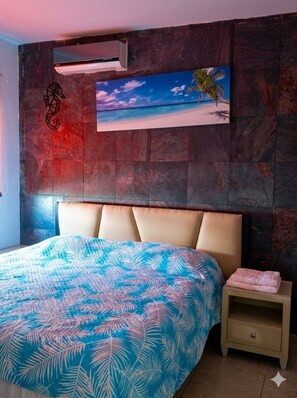A bedroom with a bed, a nightstand, a wall-mounted air conditioner, and a painting of a beach scene.