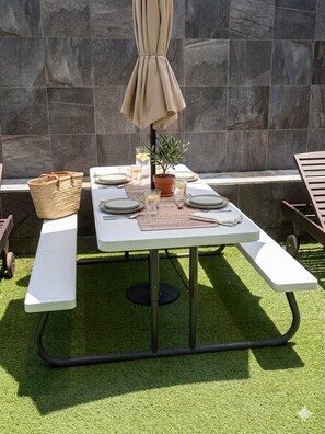 A patio table set with plates, glasses, and a basket, surrounded by artificial grass and a stone wall.
