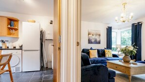 Eine moderne Küche mit Geschirrspüler, Kühlschrank und Holzschränken. Ein dunkelblaues Sofa mit Kissen und eine Blumenvase stehen auf einem Holztischchen. Eine Pendelleuchte hängt von der Decke.