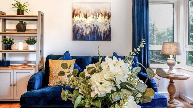Ein Wohnzimmer mit einem blauen Sofa, einer dekorativen Vase mit Blumen, einem Bücherregal und einem Gemälde an der Wand.