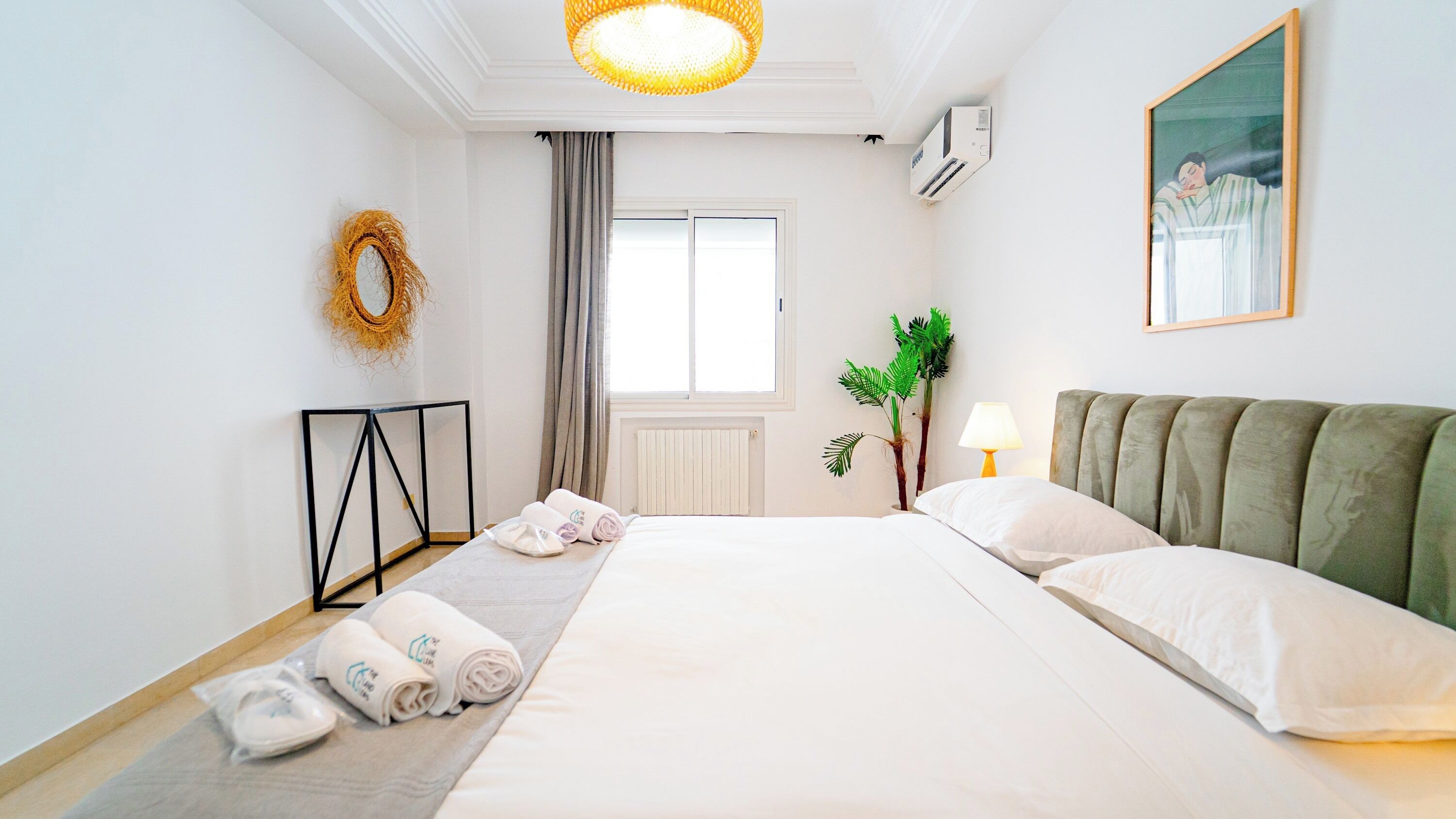 A hotel room with a bed, a standing lamp, a plant, a radiator, and a window with curtains.