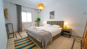A hotel room with a bed, two bedside lamps, a radiator, a window with curtains, a plant, and a painting on the wall. - Havre de Paix Apt en Plein Centre de la Marsa (La Marsa)