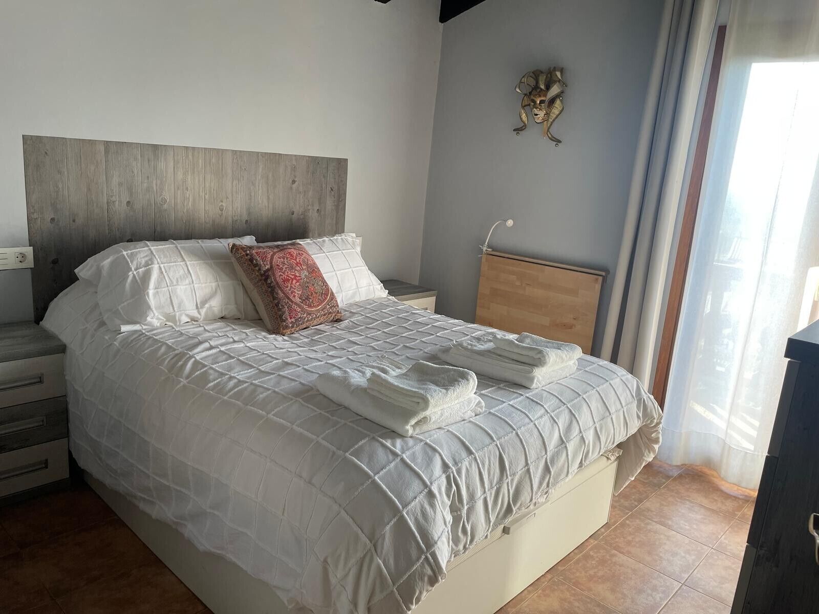 Ein ordentlich bezogenes Bett mit weißer, gesteppter Decke, einem roten Kissen mit Muster und einem Holz-Kopfteil.