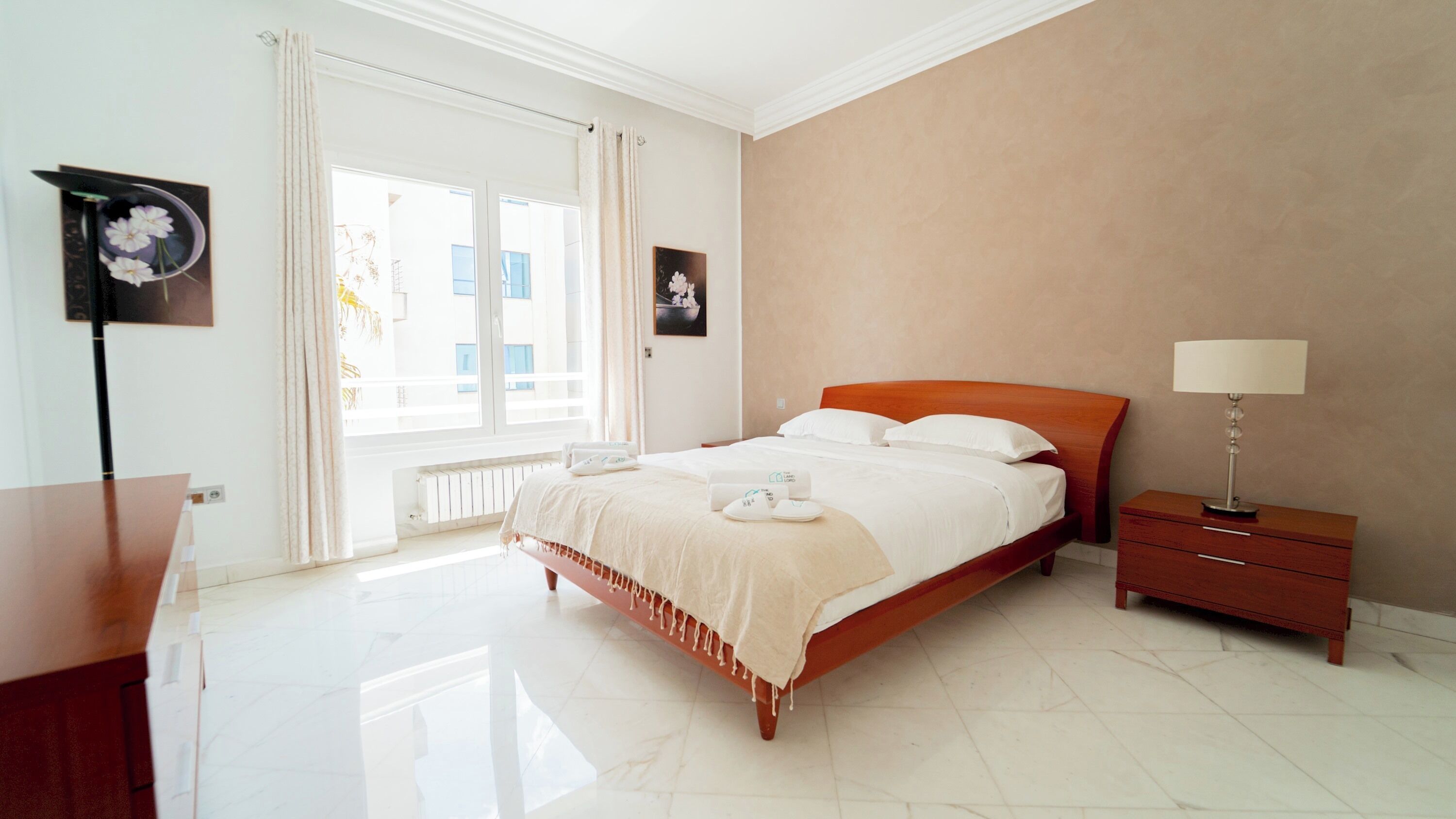 A hotel room with a large bed, a bedside table with a lamp, a view of the outside through large windows, and a framed picture on the wall.