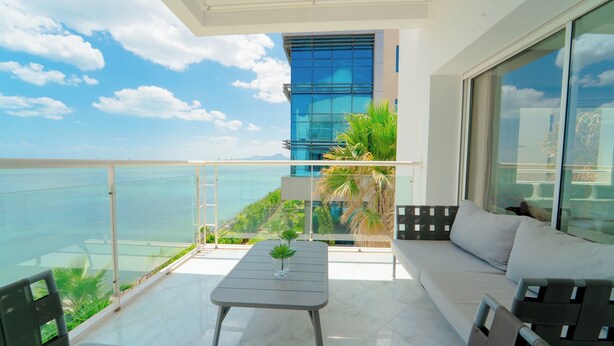 A balcony with a sofa and a table, overlooking a body of water and a modern building.