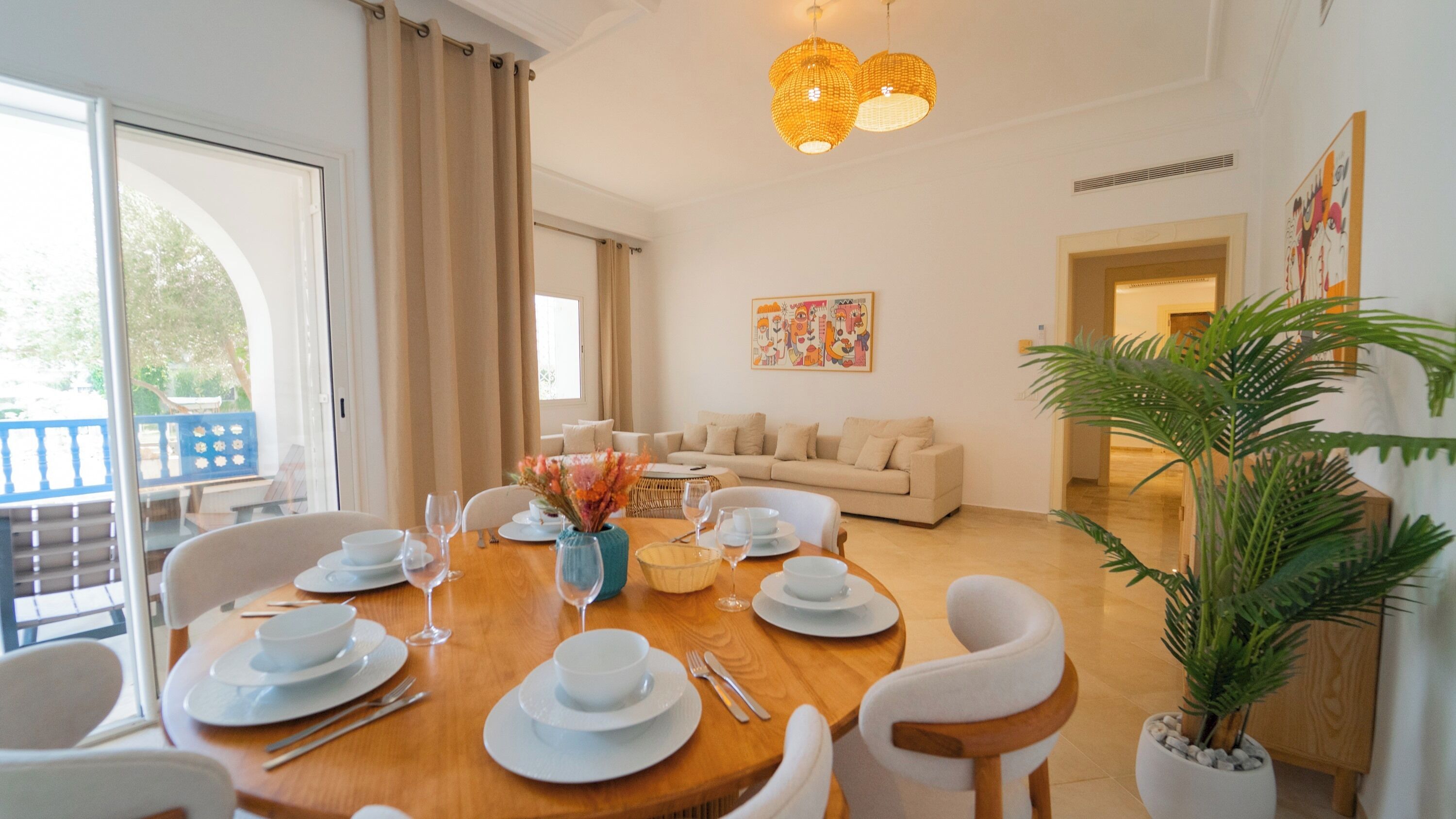 A dining area with a round table set for six, a sofa in the background, and a large window with curtains.