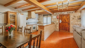 A rustic kitchen with wooden beams, a dining table with chairs, a sofa, and a stone wall.