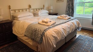 A neatly made bed with white linens and a grey quilt, a bedside table with a lamp, a window with a view of greenery, and a framed picture on the wall.