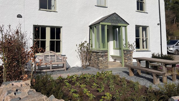 A white building with a green door and windows, a stone staircase, and a garden area with plants and a wooden bench.