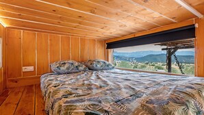 Un dormitorio con techo de madera, una cama con edredón estampado con motivos de hojas y una ventana con vistas a las montañas.