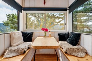 Un rincón acogedor con una mesa de madera y dos bancos acolchados, un florero con flores, y una ventana con vistas a árboles y colinas.