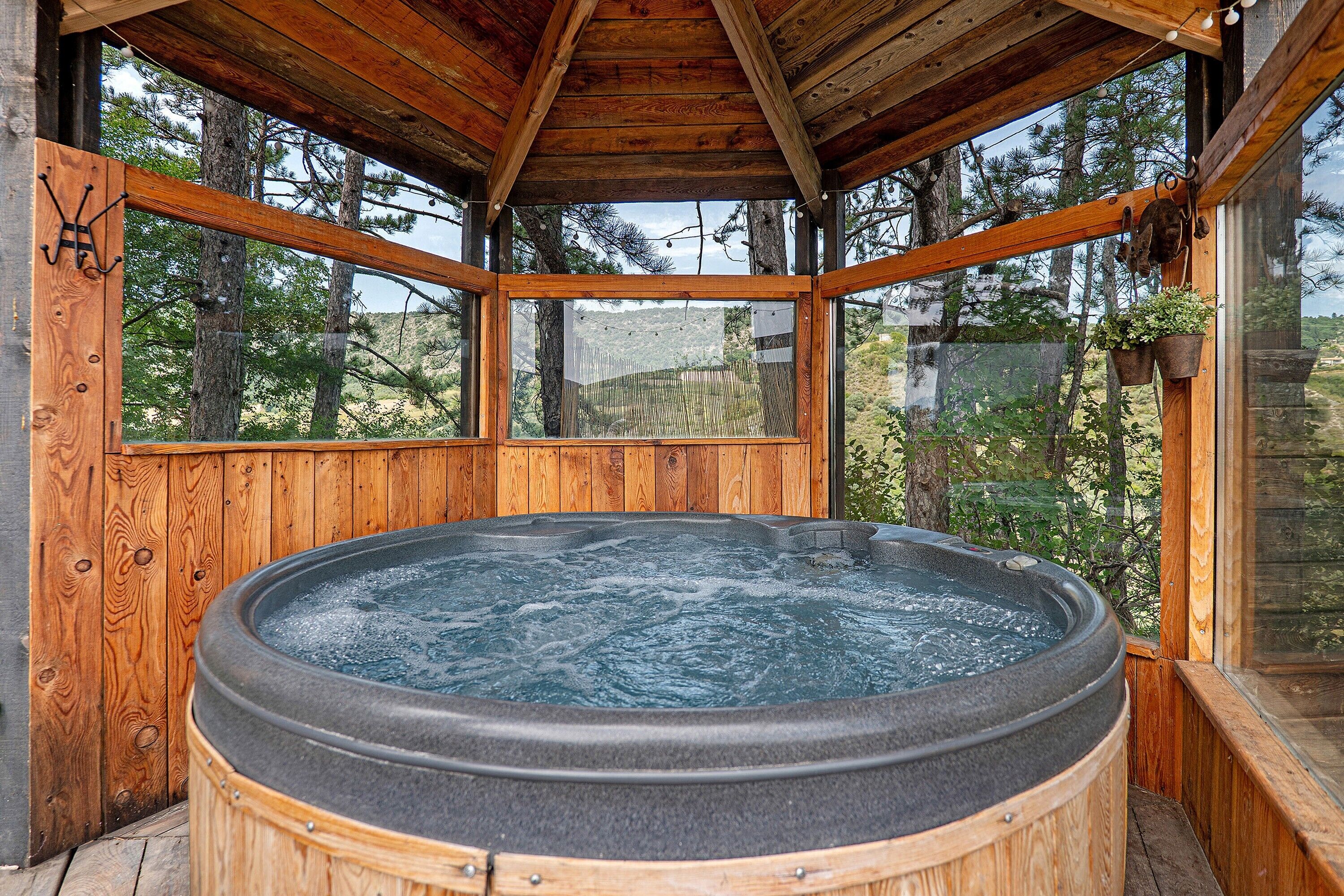A wooden cabin with a hot tub, large windows, and a view of the surrounding forest.