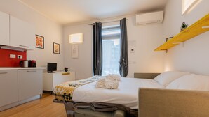 Habitación de hotel compacta con cama, escritorio, silla, kitchenette pequeña y ventana con cortinas.