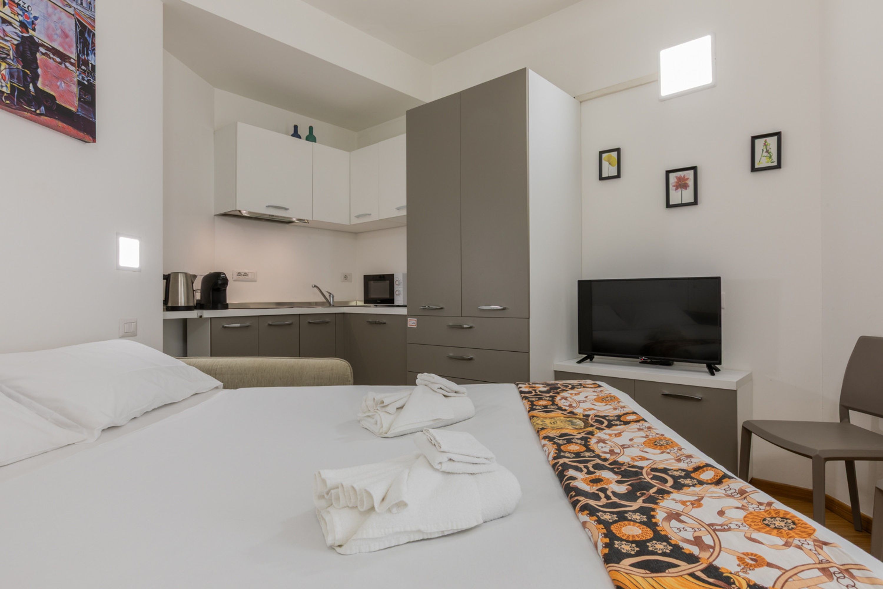 Quarto de hotel com cama, televisão, escrivaninha, cadeira, cozinha e banheiro.