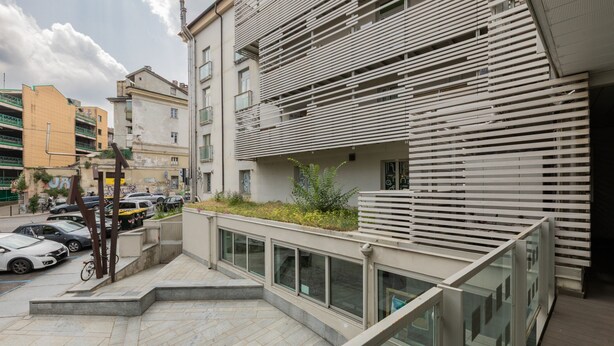 Un edificio moderno con techo inclinado, un balcón con barandilla metálica y un estacionamiento con coches y bicicletas.