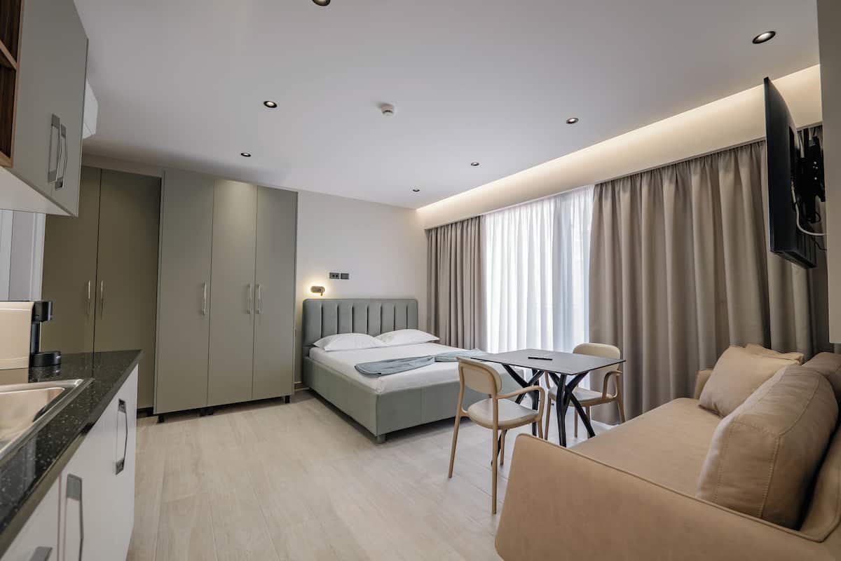 A modern hotel room with a bed, a sofa, a dining table, and a TV mounted on the wall.