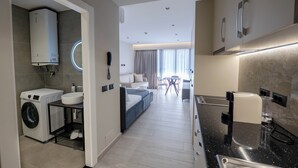 A modern hotel room with a washing machine, a sink, a bed, a dining area, and a kitchen with a coffee machine. - Blas Residence (Sarande)