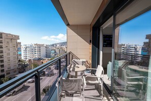 Un balcón con mesa y sillas, baranda de cristal y vista panorámica de la ciudad.