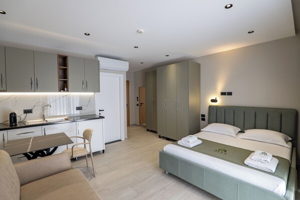Una habitación de hotel moderna con kitchenette, una cama con cabecera verde, un sofá y una zona de comedor.