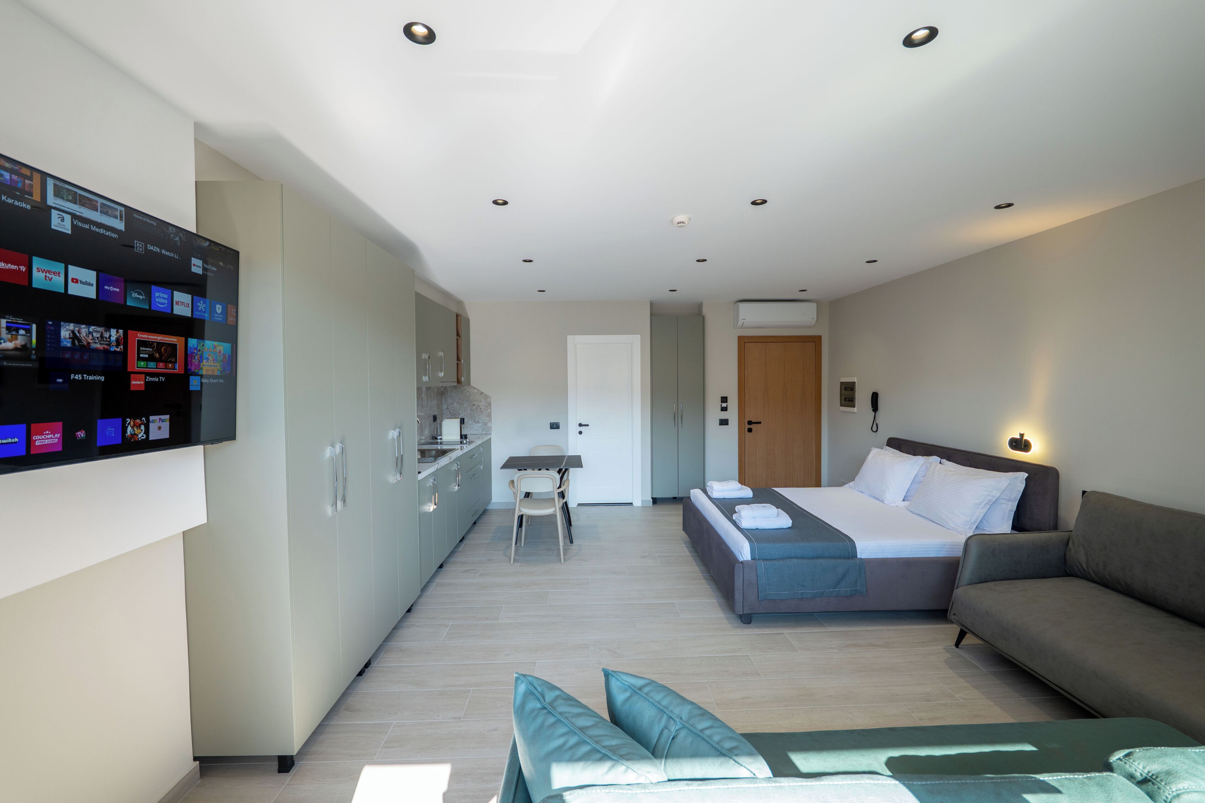 A modern hotel room with a bed, a sofa, a TV mounted on the wall, and a kitchenette.