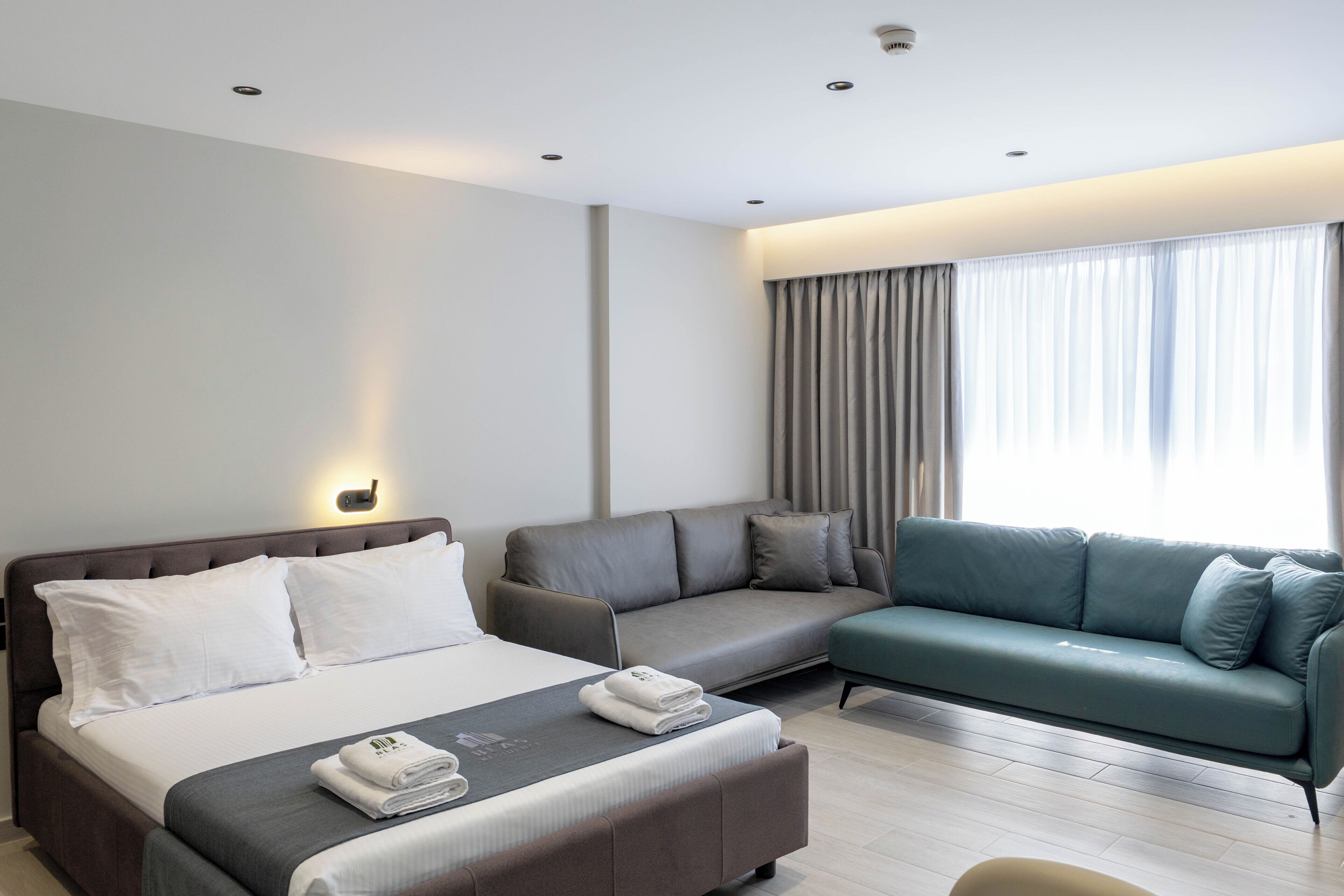 A modern hotel room with a bed, a sofa, a small ottoman, and a window with curtains.