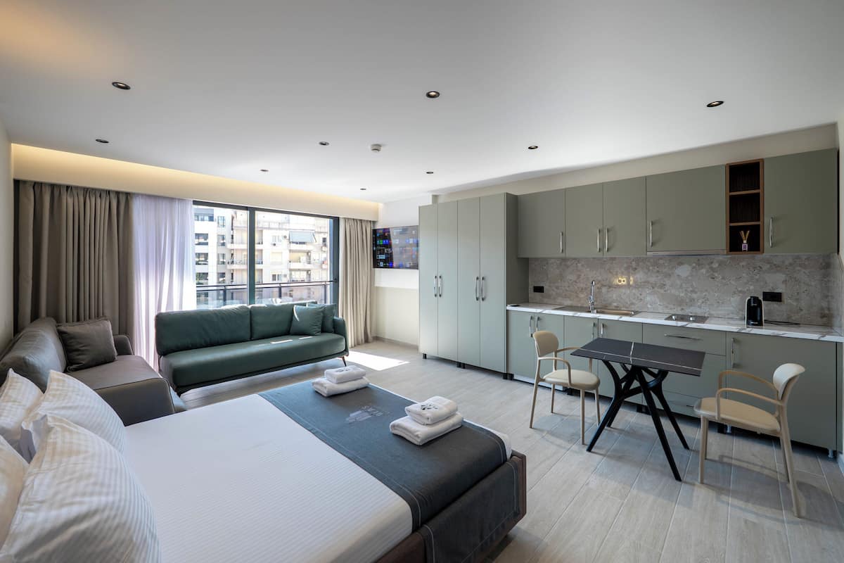 A modern hotel room with a large bed, a sofa, a dining table, and a kitchen area.