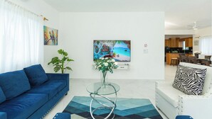 Een woonkamer met een blauwe bank, een glazen salontafel, een witte fauteuil en een kussen met een zebraprint. Aan de muur hangt een schilderij en de televisie toont een strandscène.