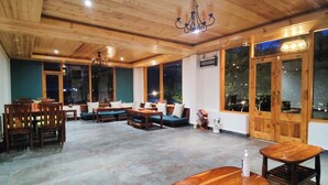 A spacious indoor area with wooden ceiling and walls, featuring a dining area with tables and chairs, a seating area with sofas, and large windows providing views outside. - NinYanWays Boutique Stay (Manali)
