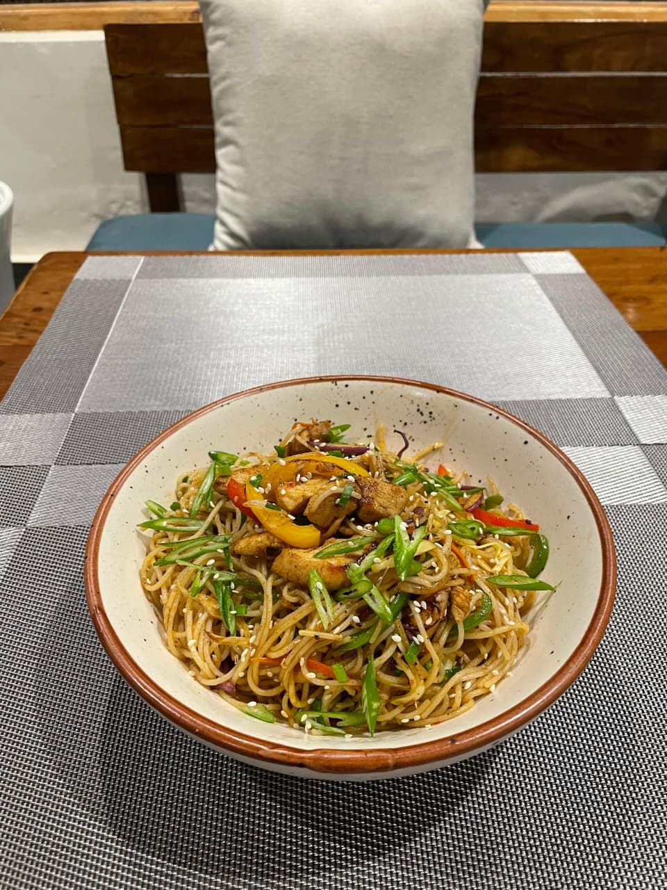 Un bol de nouilles sautées aux légumes et au poulet, posé sur une table recouverte d’une nappe à carreaux.
