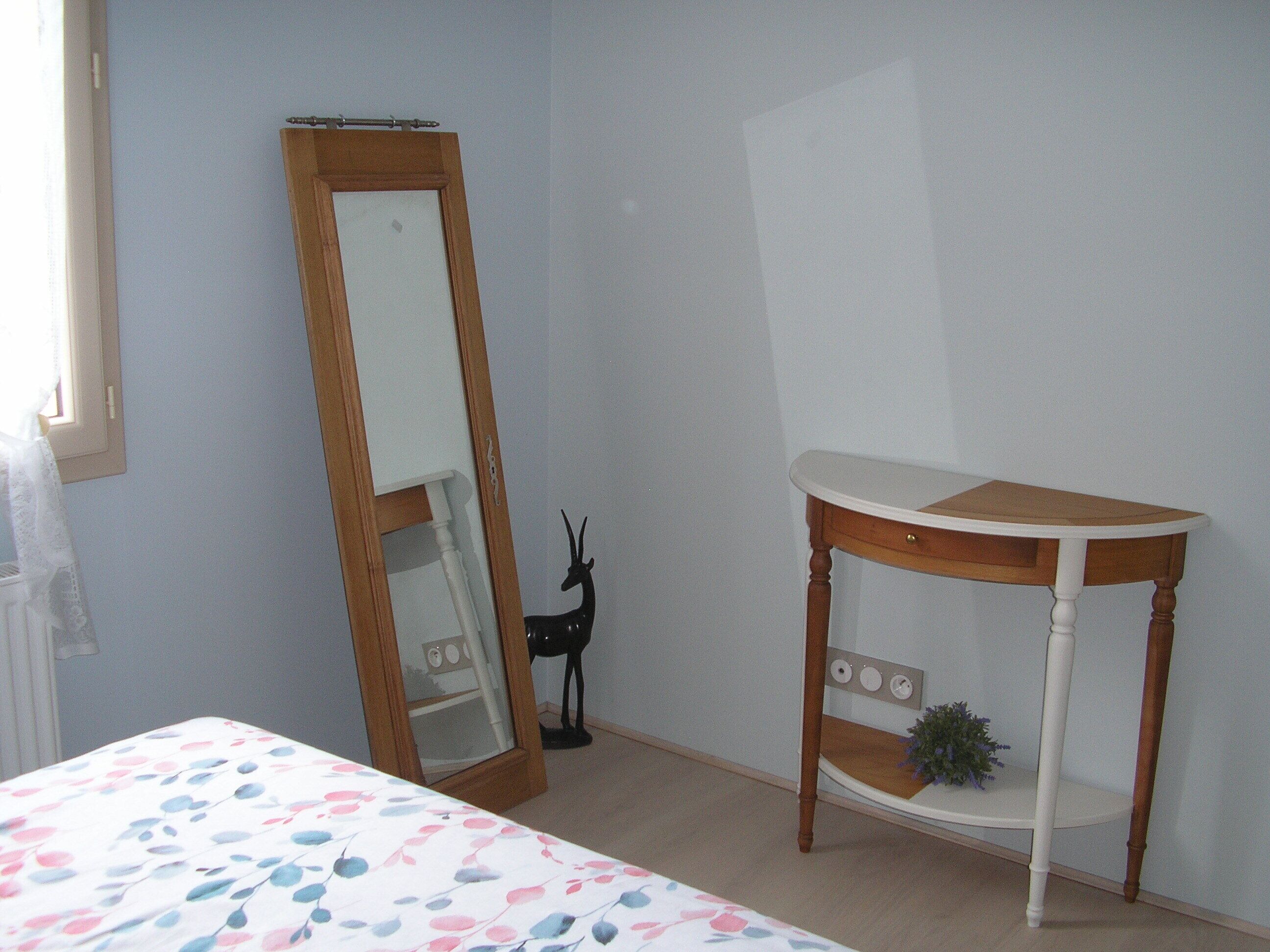 Quarto com cama, mesa de cabeceira com espelho e pequeno armário com planta.