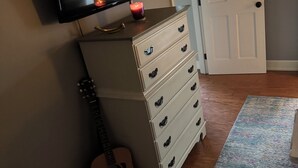 A room with a television mounted on the wall, a guitar leaning against the wall, a dresser with multiple drawers, and a door in the background.