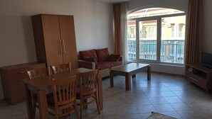 A furnished living room with a dining table, chairs, a sofa, a TV stand, and a large window with a view of buildings.