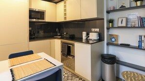 A compact kitchen with a microwave, toaster, and sink. There is a small dining table with a placemat. A trash bin and a shelf with books and a plant are also visible. - Charming  studio in the 17th, ideal for 2 (Paris)