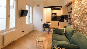 A compact living space with a kitchenette, a green sofa, a small round table, and a stone wall. - Charming  studio in the 17th, ideal for 2 (Paris)