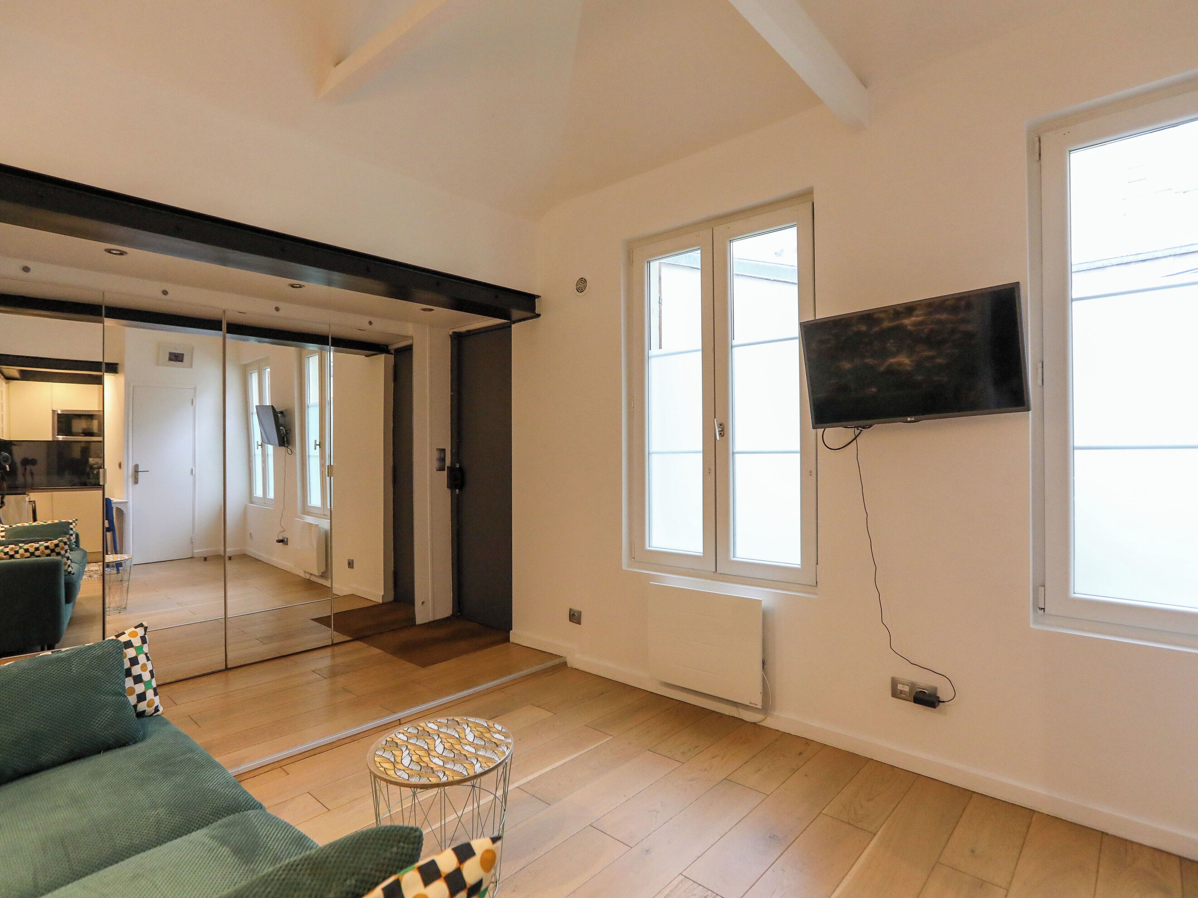 Een moderne woonkamer met een bank, een klein rond tafeltje, een aan de muur bevestigde televisie en een groot raam.