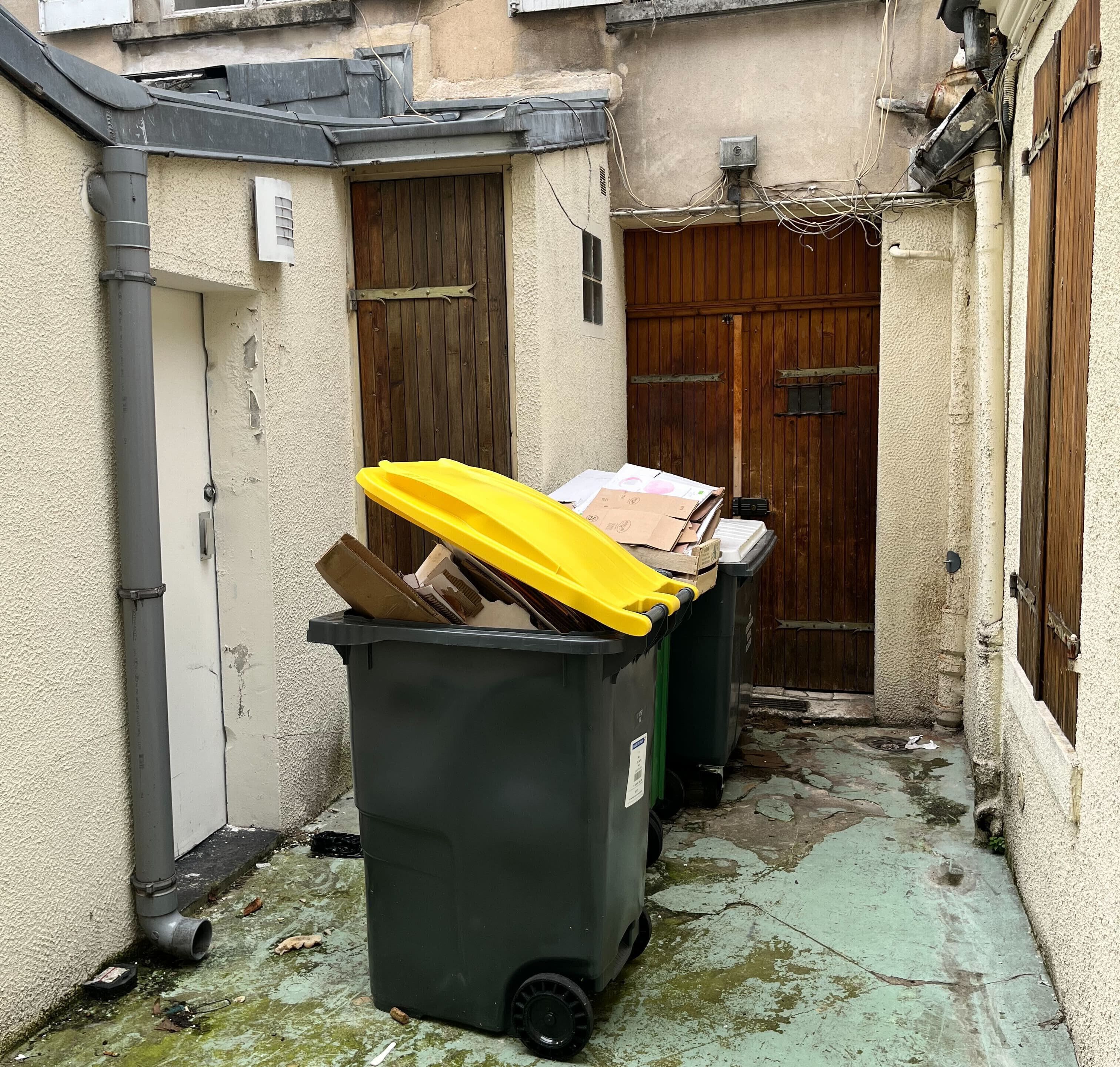 A narrow alley with two black dumpsters, one with a yellow lid, and a wooden door in the background.
