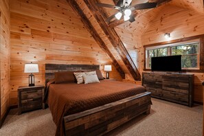 A wooden cabin bedroom with a bed, two bedside tables, a dresser, a ceiling fan, and a TV.